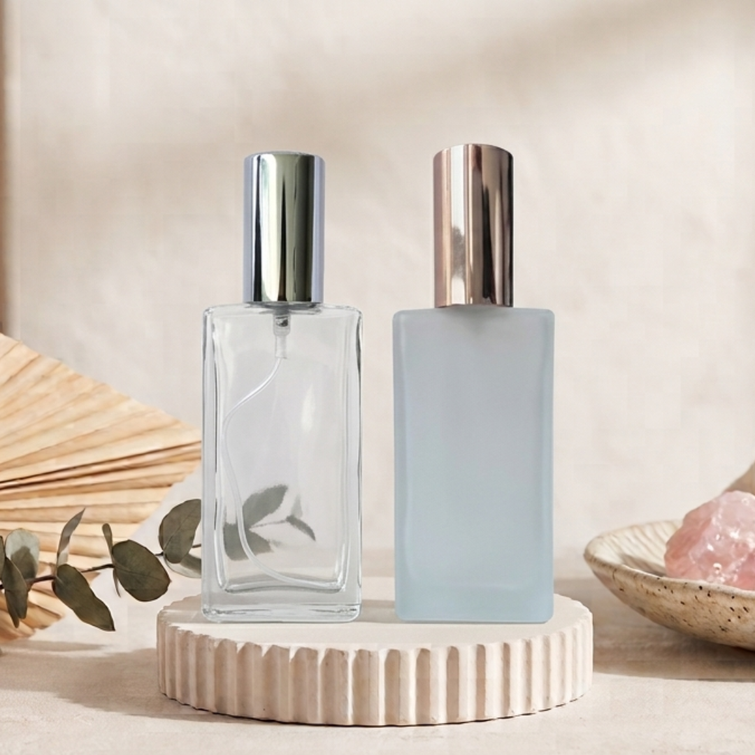 Two rectangular perfume bottles displayed side by side on a ribbed beige pedestal. The left bottle is made of transparent glass with a reflective silver cap, and the right bottle is frosted in a pale icy blue with a metallic rose-gold cap.